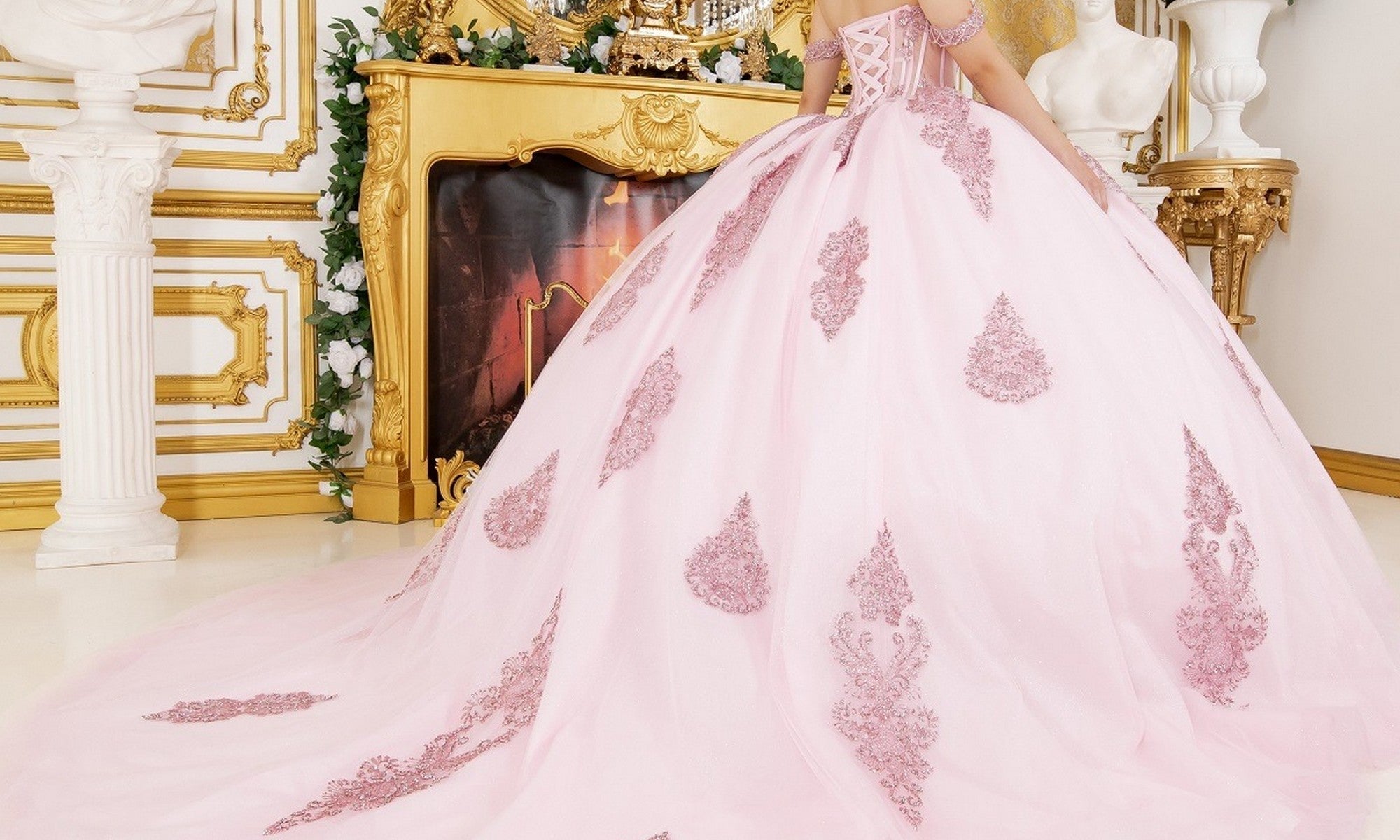 A person wearing the Quinceanera Dress 8190J by Cinderella Couture, featuring delicate lace details in a pastel hue, stands in front of an ornate gold and white fireplace in an elegant room.